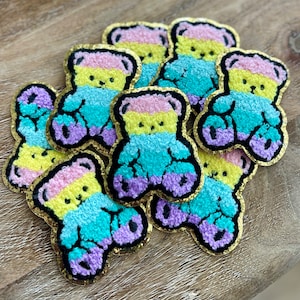 May include: A collection of colorful teddy bear patches. Each patch features a rainbow design with pink, yellow, teal, and purple sections, outlined in black and edged with gold. The patches are made of a textured material.