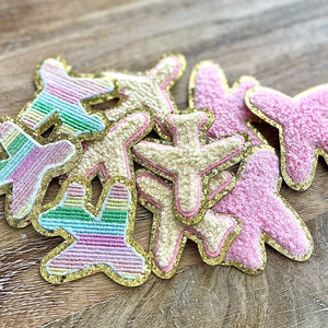 May include: A collection of airplane-shaped patches in various colors. Some are pink, cream, and rainbow-striped, with gold glitter edging. The patches have a textured, plush appearance, suggesting an embroidered or appliquéd design.