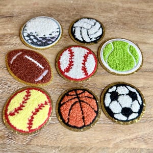 May include: A collection of eight circular and oval embroidered patches featuring various sports balls, including a golf ball, volleyball, football, baseball, tennis ball, softball, basketball, and soccer ball, each with a gold glitter border.