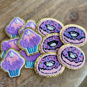 May include: A collection of embroidered patches featuring cupcakes and donuts. The cupcakes have purple frosting with pink accents and blue liners. The donuts are pink with purple frosting and colorful sprinkles. Each patch has a gold glitter border.