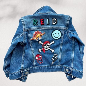 May include: A blue denim jacket with patches. Patches include a dinosaur, a smiley face, a skull and crossbones, a Spider-Man graphic, a lightning bolt, and the name "REID" in colorful letters.