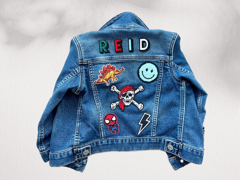May include: A blue denim jacket with patches. Patches include a dinosaur, a smiley face, a skull and crossbones, a Spider-Man graphic, a lightning bolt, and the name "REID" in colorful letters.