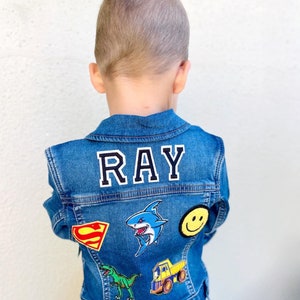 May include: A blue denim jacket with the name "RAY" in white block letters on the back. The jacket is decorated with colorful patches, including a shark, a dinosaur, a smiley face, a Superman logo, and a dump truck.