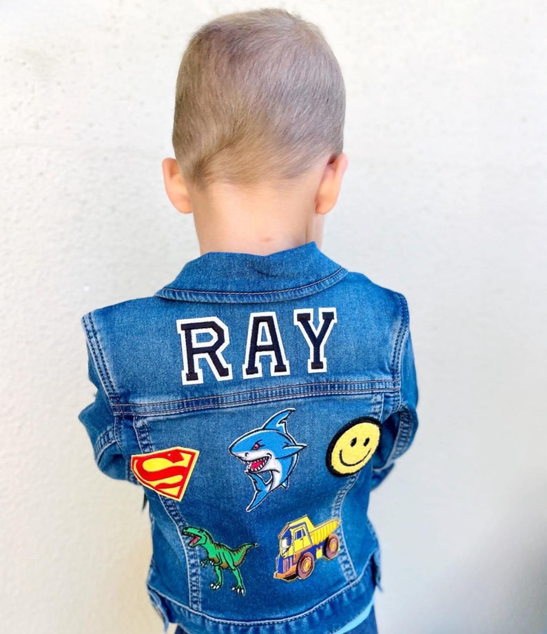 May include: A blue denim jacket with the name "RAY" in white block letters on the back. The jacket is decorated with colorful patches, including a shark, a dinosaur, a smiley face, a Superman logo, and a dump truck.