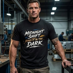 May include: Black t-shirt with the text "MIDNIGHT SHIFT DOES IT IN THE DARK" in white, blue, and gold. The shirt is worn by a person in a factory setting, with a tattoo on the arm. The person is wearing brown pants.