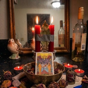 May include: Two red candles joined by gray tape with a bay leaf between them stand behind a bowl containing a tarot card depicting an angel and two figures. The scene includes scattered dried roses, small red tea light candles, a wax seal stamp, a blue pen, and a small metal birdcage.