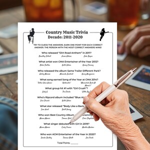 May include: A white sheet of paper with a country music trivia game printed on it. The trivia game is titled "Country Music Trivia Decade: 2011-2020." A person is holding a pen over the paper, ready to write.