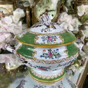 May include: A ceramic serving dish with a lid. The dish has a white base with green and gold accents, and is decorated with floral patterns in pink, blue, and yellow. It has two handles and a decorative knob on the lid. The dish is likely for serving food.