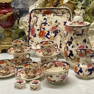 Op de afbeelding: Een theeservies van porselein met een crèmekleurige basis en ingewikkelde bloemmotieven in oranje, roze en blauw, inclusief een theepot met deksel, een melkkan, een suikerpot, vier kopjes met schoteltjes, een serveerblad en twee kleine doosjes.