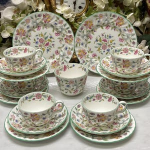 May include: A set of fine china featuring a floral pattern with pink, yellow, and blue blossoms on a white background. The collection includes two large plates, five teacups, and five saucers, all finished with a delicate green rim detail.