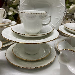 Peut inclure: Un ensemble de tasses à thé et soucoupes en porcelaine blanche avec des bords festonnés et un fin liseré doré. Le service comprend des tasses empilées sur des soucoupes, un grand plat carré et un pot à lait, tous avec un motif en relief.
