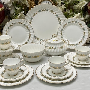Op de afbeelding: Een wit porseleinen servies met een geschulpte rand en een delicaat patroon van groene en gouden bladeren. De collectie bevat een grote serveerschaal, een suikerpot met deksel, een kom en diverse theekopjes met bijpassende schoteltjes.