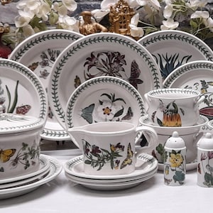 May include: A collection of white ceramic dinnerware featuring botanical illustrations of flowers and butterflies with green leaf-patterned borders, including plates, a lidded tureen, a creamer, bowls, and salt and pepper shakers.
