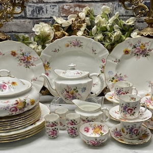 May include: A porcelain dinnerware set featuring a cream-coloured base with scalloped edges and a colourful floral pattern of pink, yellow, and blue flowers. The collection includes plates, a lidded tureen, a teapot, a creamer, cups, and saucers.