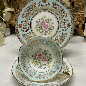 Op de afbeelding: Een lichtblauw porseleinen theekop en schotel set met een bijpassend decoratief bord. De stukken zijn voorzien van gouden filigraan, een centraal bloemenboeket met roze bloemen en het koninklijk monogram ER met een kroon in gouden en rode letters.