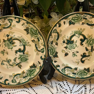 Puede incluir: Dos platos de cerámica blanca con un diseño floral verde y dorado. Los platos tienen un borde dorado y están en soportes de exhibición.