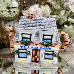 Op de afbeelding: Een keramische theepot in de vorm van een gebouw met twee verdiepingen met een grijs dak, een bakstenen gevel en blauwe ramen. De theepot heeft een bord met de tekst "VILLAGE BANK" en een bruine deur.