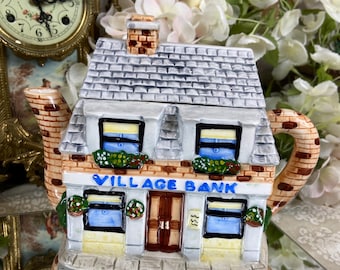 May include: A ceramic teapot shaped like a two-storey building with a grey roof, brick facade, and blue windows. The teapot has a sign that reads "VILLAGE BANK" and a brown door.