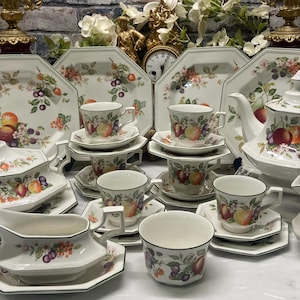 Op de afbeelding: Een wit porseleinen servies met een kleurrijk patroon van fruit en bessen met groene bladeren. De collectie bevat achthoekige borden, theekoppen met schotels, een theepot, een sauskom, een melkkan en een serveerschaal met deksel en handvatten.