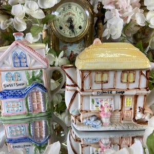 May include: Two ceramic teapots shaped like houses. The teapot on the left is painted to look like a sweet shop with the words "Sweet Shop" on the front. The teapot on the right is painted to look like a flower shop with the words "Flower Shop" on the front.