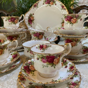 Può includere: Un set di tazze da tè e piattini vintage con un motivo floreale. Le tazze e i piattini sono bianchi con un bordo dorato e presentano un motivo floreale rosso, rosa e giallo. Il set comprende una teiera, un bricco per la panna e una zuccheriera.