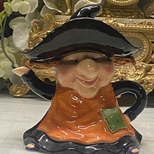 Könnte beinhalten: Eine Keramik-Teekanne in Form einer Hexe mit schwarzem Spitzhut, schwarzem Langarm-Oberteil und einer orangefarbenen Schürze mit einem kleinen grünen quadratischen Flicken. Die Figur hat eine große Nase, ein Lächeln und sichtbare lila Schuhe.