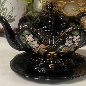 Könnte beinhalten: Eine schwarze Keramik-Teekanne mit passender Untertasse, glänzendem Finish, goldfarbenen Akzenten und handgemalten Blumenmotiven in Weiß, Rosa und Grün. Die Teekanne verfügt über einen geschwungenen Griff, einen Ausguss und einen verzierten Deckel.