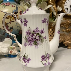 Puede incluir: Cafetera de porcelana blanca con tapa, decorada con motivos florales de violetas moradas. La cafetera tiene un borde dorado y un asa curva. El diseño es de estilo clásico y vintage.