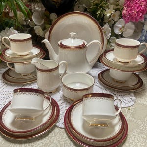 May include: A white tea set with gold trim and red accents. The set includes a teapot, creamer, sugar bowl, and four teacups and saucers.