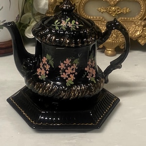 Op de afbeelding: Een zwarte keramische theepot met een bijpassende zeshoekige basis. De theepot heeft gouden randen en delicate roze bloemmotieven. De handgreep is gebogen en het deksel is bekroond met een gouden knop. Een decoratief item.