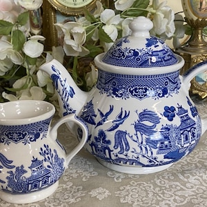 May include: A white porcelain teapot with a lid and a matching creamer, both featuring a blue Willow pattern. The teapot has a curved spout and handle. The creamer has a small handle and spout. The set is on a patterned surface.