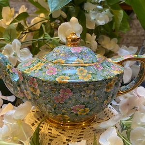 Könnte beinhalten: Eine blaue und weiße Blumenvase mit goldenen Verzierungen. Die Teekanne hat ein Blumenmuster mit rosa, gelben und grünen Blumen. Die Teekanne steht auf einer weißen Häkeldeckchen.