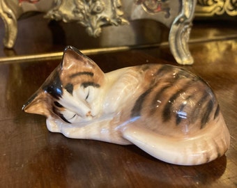 Puede incluir: Una figurita de porcelana de un gato atigrado durmiendo con marcas marrones, negras y blancas. El gato está enroscado en una posición para dormir con la cabeza apoyada en las patas.