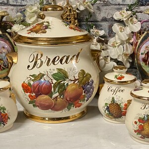 Puede incluir: Un juego de cocina de cerámica color crema que incluye un tarro grande para pan, tres botes pequeños etiquetados como Sugar, Coffee y Tea, y dos jarrones ornamentados. Cada pieza está decorada con bordes dorados e ilustraciones de frutas.