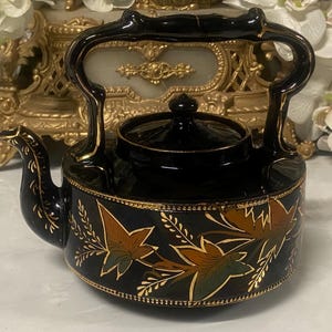 Op de afbeelding: Zwarte keramische theepot met een handvat en tuit, met gouden randen en bladeren in bruin- en groentinten. De theepot heeft een deksel en een decoratieve gouden rand.