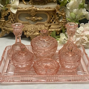 Peut inclure: Un ensemble de toilette en verre pressé rose comprenant un plateau rectangulaire, deux bougeoirs, deux pots avec couvercle et une petite coupelle. Chaque pièce présente un motif géométrique en étoile et en losange en verre rose translucide.