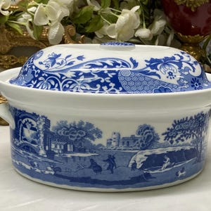 Puede incluir: Una fuente de cerámica blanca con tapa, decorada con un patrón azul de transferencia que representa un paisaje pastoral con figuras, edificios y árboles. El recipiente redondo tiene dos pequeñas asas laterales y una tapa abovedada a juego.