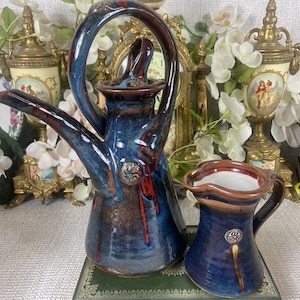 Puede incluir: Un juego de cafetera y crema de cerámica. La cafetera es azul con detalles marrones y un asa única. La crema es azul con borde y asa marrones. Ambas piezas tienen detalles decorativos y un acabado brillante.