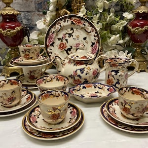 Op de afbeelding: Een theeservies van porselein met een crèmekleurige basis en ingewikkelde bloemmotieven in oranje, roze en donkerblauw. De set bevat een theepot met deksel, een melkkan, een serveerschaal, een klein kommetje en diverse kopjes met bijpassende schoteltjes.