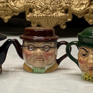 May include: Three ceramic teapots with human face designs. The teapots have black, brown, and green hats. The faces are painted with details like beards, glasses, and winks. The teapots have brown handles and spouts.