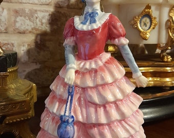 以下が含まれることがあります： ピンクと赤のドレスを着た女性の磁器製フィギュア。青いハンドバッグを持っています。彼女は青と白の帽子をかぶり、白いレースの襟をしています。