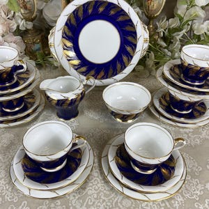 Può includere: Un servizio da tè vintage con tazze, piattini e un piatto da portata in porcellana bianca. Il set presenta un design a foglie blu navy e oro con bordi dorati. Include una lattiera e una piccola ciotola. Le tazze sono impilate.