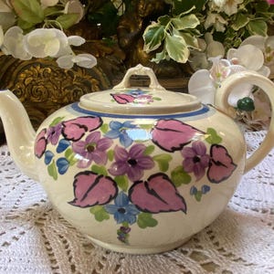 May include: A cream-coloured teapot with a floral design. The teapot has a pink, blue, and green floral pattern with pink leaves. The teapot has a lid and a handle.