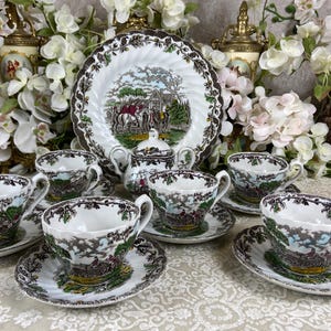 Puede incluir: Un juego de té vintage con seis tazas y platillos, un plato grande y un azucarero. El juego de porcelana blanca tiene un diseño de paisaje marrón y verde con un borde decorativo. El juego está dispuesto sobre un mantel blanco.