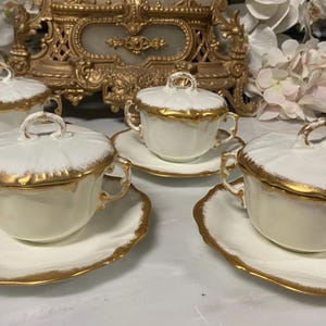 Op de afbeelding: Een set van vier witte porseleinen kommen met deksel, voorzien van twee zijhandvaten en bijpassende schoteltjes. Elk stuk heeft een geschulpte rand en een decoratieve gouden afwerking langs de randen, de deksels en de handvaten.