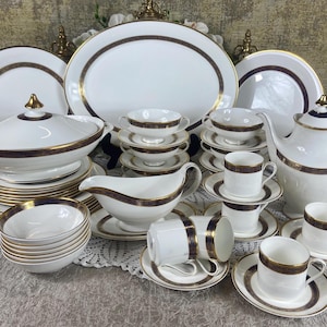 Puede incluir: Un juego de vajilla de porcelana blanca con ribetes dorados y azules. El juego incluye platos, cuencos, tazas, platillos y una tetera.