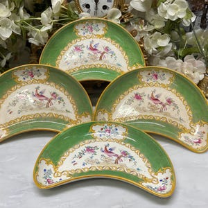 May include: A set of four crescent-shaped ceramic dishes featuring a green border with a textured pattern, gold trim, and a central illustration of a colourful bird perched on a branch surrounded by floral motifs on a white background.