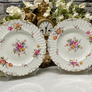 May include: Two white porcelain plates featuring a central floral bouquet of pink, yellow, and purple flowers. The plates have a fluted rim design with gold-painted scalloped edges and additional small floral clusters scattered around the border.