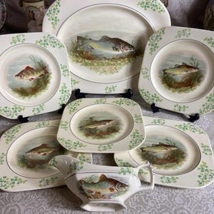 Può includere: Una collezione di piatti vintage color crema e una salsiera, ognuno con un'illustrazione dettagliata di un pesce in un ambiente naturale. I piatti hanno una forma quadrata con angoli arrotondati e sono decorati con accenti floreali verdi.