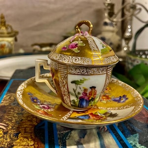 Puede incluir: Un juego de taza y platillo de porcelana vintage con borde y detalles dorados. La taza tiene tapa y asa. El juego presenta una escena pintada de personas en un jardín, con detalles florales y dorados.